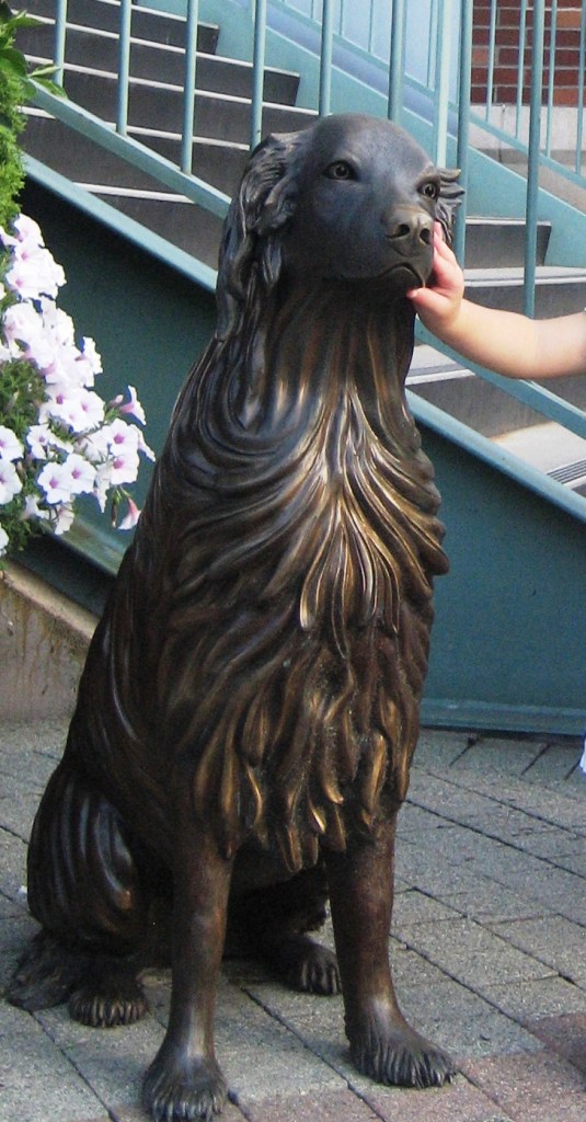 Bronze statue of a dog sitting in front of some fresh flowers, a child's small hand on the dog's face.