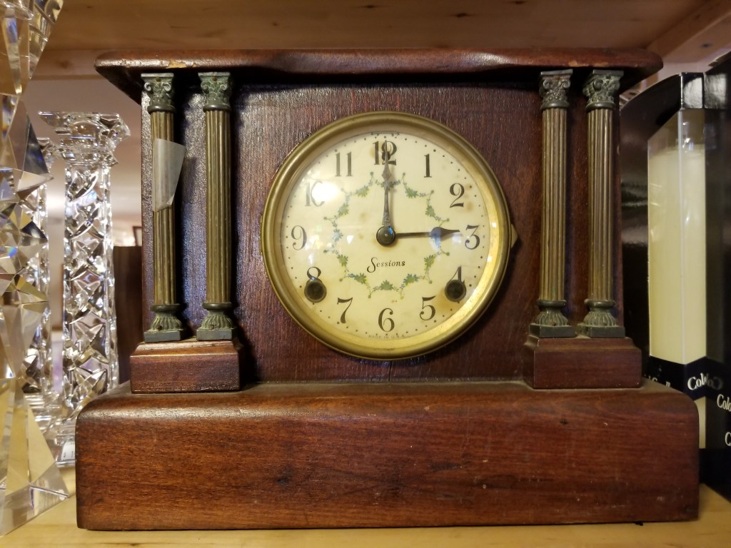Wooden clock with four columns, two on either side of the clock face - hands stuck at 12 and 3 for 3 o'clock. A crystal candle holder can be seen on the left side of the clock.
