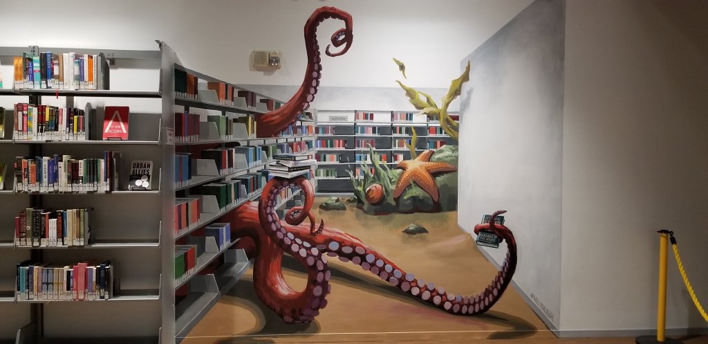 Mural on the wall of the Seattle Public Library that gives the illusion of shelves continuing into the distance. A sea creature stretches out its tentacles and grabs books to read. A giant starfish and sea kelp are in the far corner. 