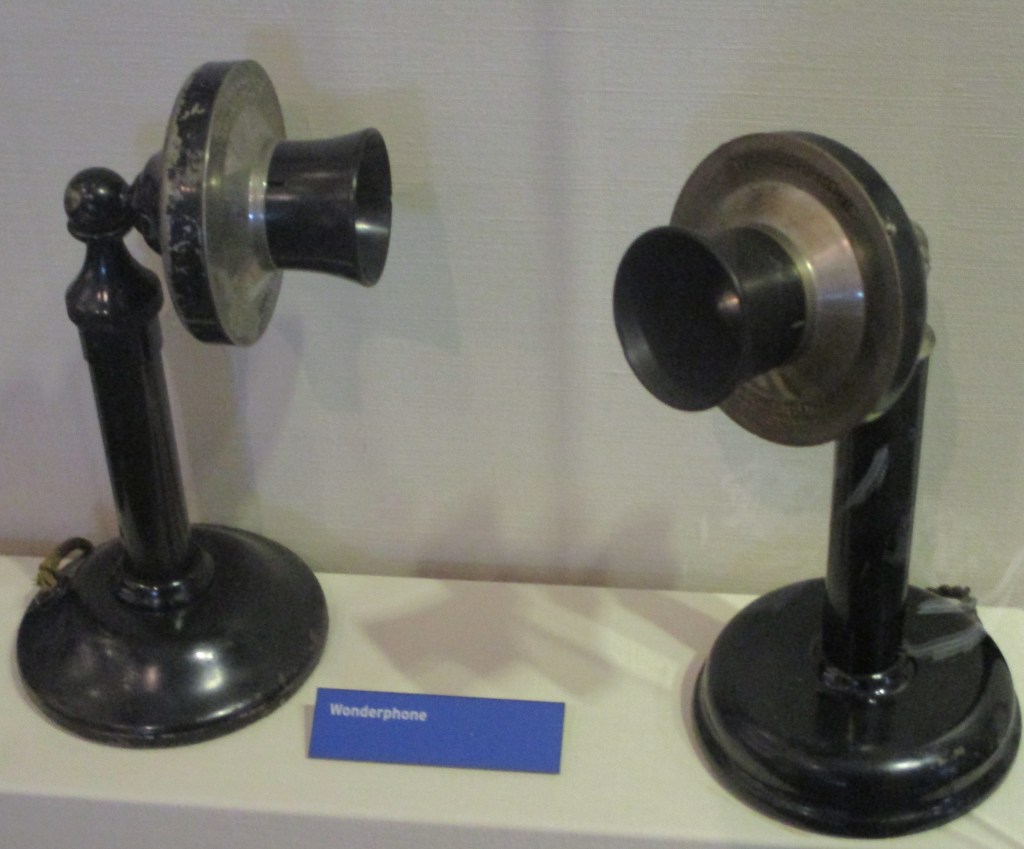 Two old-fashioned phones with a wide-mouth speaker/listening piece atop a skinny pipe, attached to a wide base. Plaque labels them the Wonderphone.