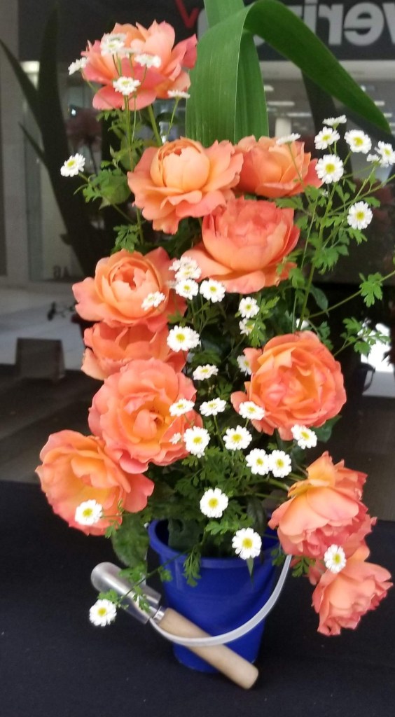 An arrangement of roses and tiny daisies in a small blue pail, with a tiny gardening shovel leaning against it.
