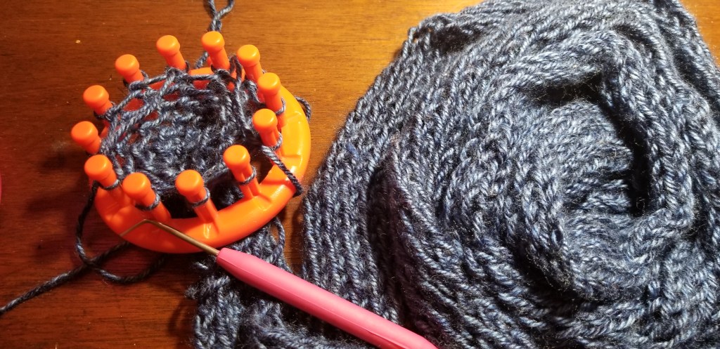 A circular knitting knobby with blue yarn wrapped around the pegs. A small hook laying in front of it. Both beside a coil of knitted blue yarn that may (or may not) become a scarf.
