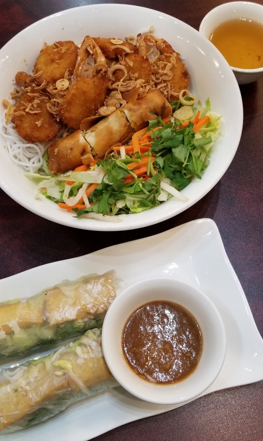 Two tofu spring rolls and peanut sauce on one plate. Above that, a bowl of rice noodles, vegetables, fried shrimp, and a cut-up fried egg roll. To the right, a small cup of fish sauce.