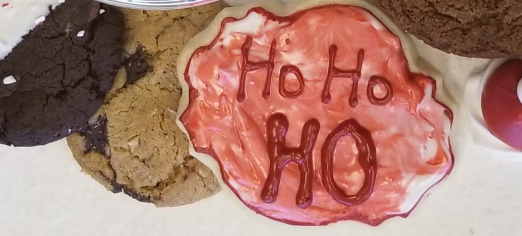 Assorted cookies on a plate. The largest cookie has red and white frosting, with the words "Ho, Ho, HO" written in red frosting.
