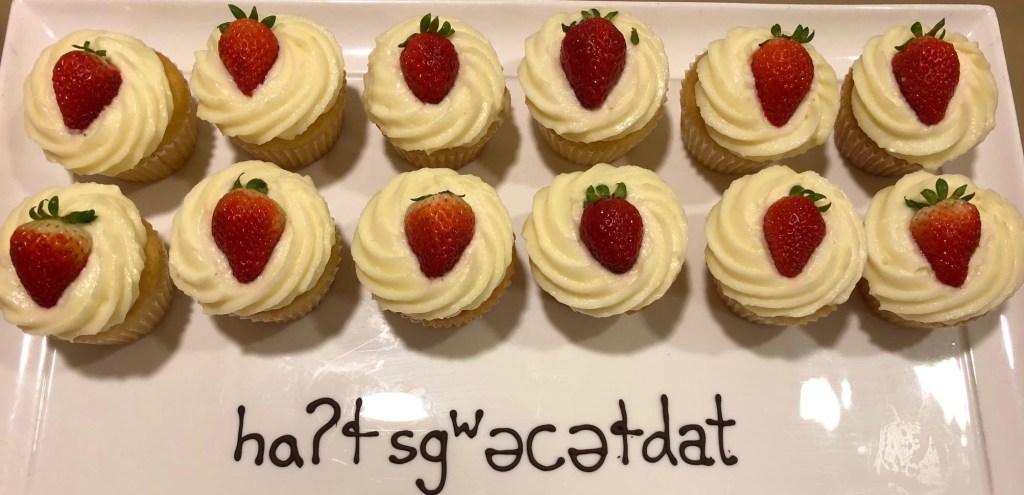 On a white tray, two rows of white-frosted cupcakes topped with strawberries. Text below, written in chocolate frosting, "Happy Birthday" in the Lushootseed language.