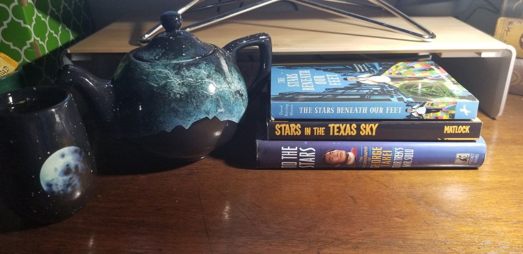 A ceramic/glazed stemless wine glass with a moon and stars painted on it.
A ceramic/glazed teapot with a mountain outline underneath a star-filled sky. 
Three books stacked atop another - from top to bottom:
- The Stars Beneath Our Feet, by David Baclay Moore
- Stars in the Texas Sky, by Stephen Matlock and
- To the Stars, by George Takei