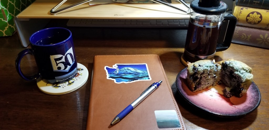 A blue coffee cup on a coaster. On it, an  open book in white, with the number "50" in the center of it and confetti falling over both. Small text in blue on light blue: "Celebrating 50 Years." In white, beneath that, "King County Library System."

Beside that, a brown faux level 6 bty8 portfolio with two stickers and a blue pen on it. First sticker is shaped like Washington State, with Mount Tahoma in the middle. Various shades of blue. Lower corner, a square sticker of Storm's art called "Ocean."

To the right of that, a red ceramic plate with a halved blueberry muffin on it. By that, a small tea press filled with tea
