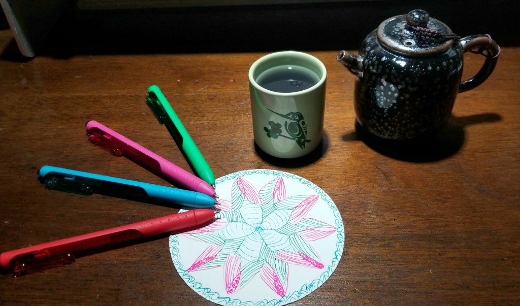 Four gel pens - red, blue, pink, and green, atop a Zentangle mandala. Beside it, a small, green teacup with a hummingbird filled with blue tea. Beside that, a small Japanese glaze teapot.