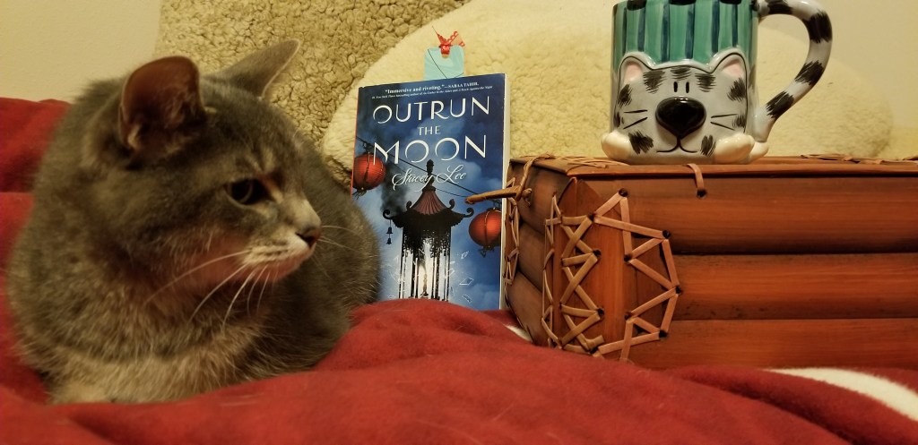 Gray and white cat on a red blanket on a bed. Propped beside it, a book titled, "Outrun the Moon" by Stacey Lee. Propped on a bamboo box, a gray and white cat in front of green stripes cup. The paws are the base, a curved tail the handle.