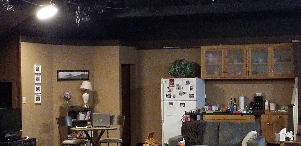 A small stage set for the play, "Tiny Beautiful Things" Stage light overhead, a collection of photos on the wall, bookcase, and lamp behind a small table with an open computer on top. Two cafe chairs facing each other.
A kitchen with a fire covered in photos, a plant up top, assorted items out and about on the kitchen counters. A couch a pillow and throw.