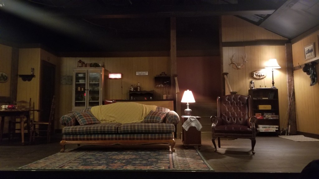 Theater stage set with a sofa, end table,  lamp,  rug,  table, bar, paintings and odds and ends of a boarding house. 