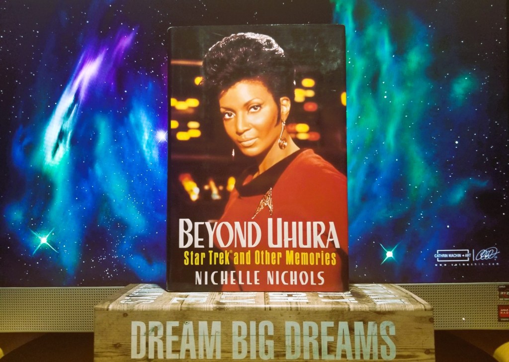 Nichelle Nichol's autobiography, "Beyond Uhura: Star Trek and Other Memories" propped atop a pencil box with the words "Dream Big Dreams" on it. Both sit in front of a cosmos painting image (digital copy) of art by Cathrin Machin