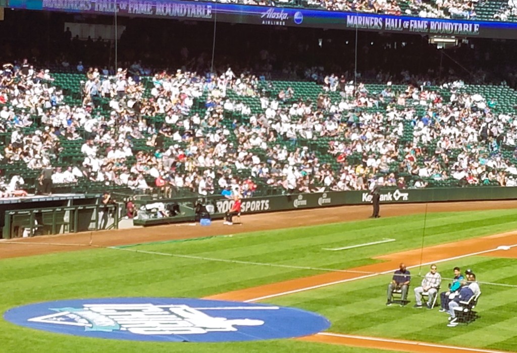 Alvin Davis, Edgar Martinez, Ichiro's translator, Ichiro, Ken Griffey Jr. and Rick Rizz sitting on folding chairs in between the pitcher's mound and home plate that is covered by a round blue banner with a silhouette of Ichiro Suzuki, the name Ichiro in all caps, and Mariners Hall of Fame.