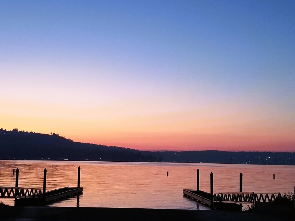 Lake Sammamish, with a boat launch site int eh center, pillars in the distance, hills with houses, and the colors of sunset.