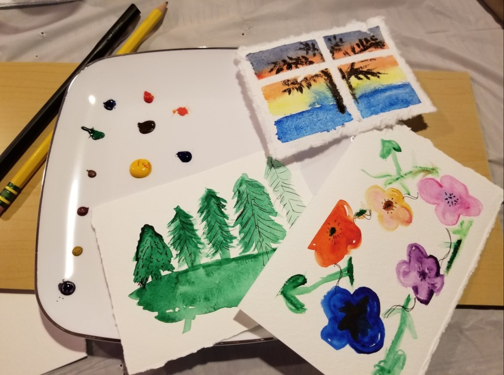 A trio of miniature watercolors beside a palate of various colors.