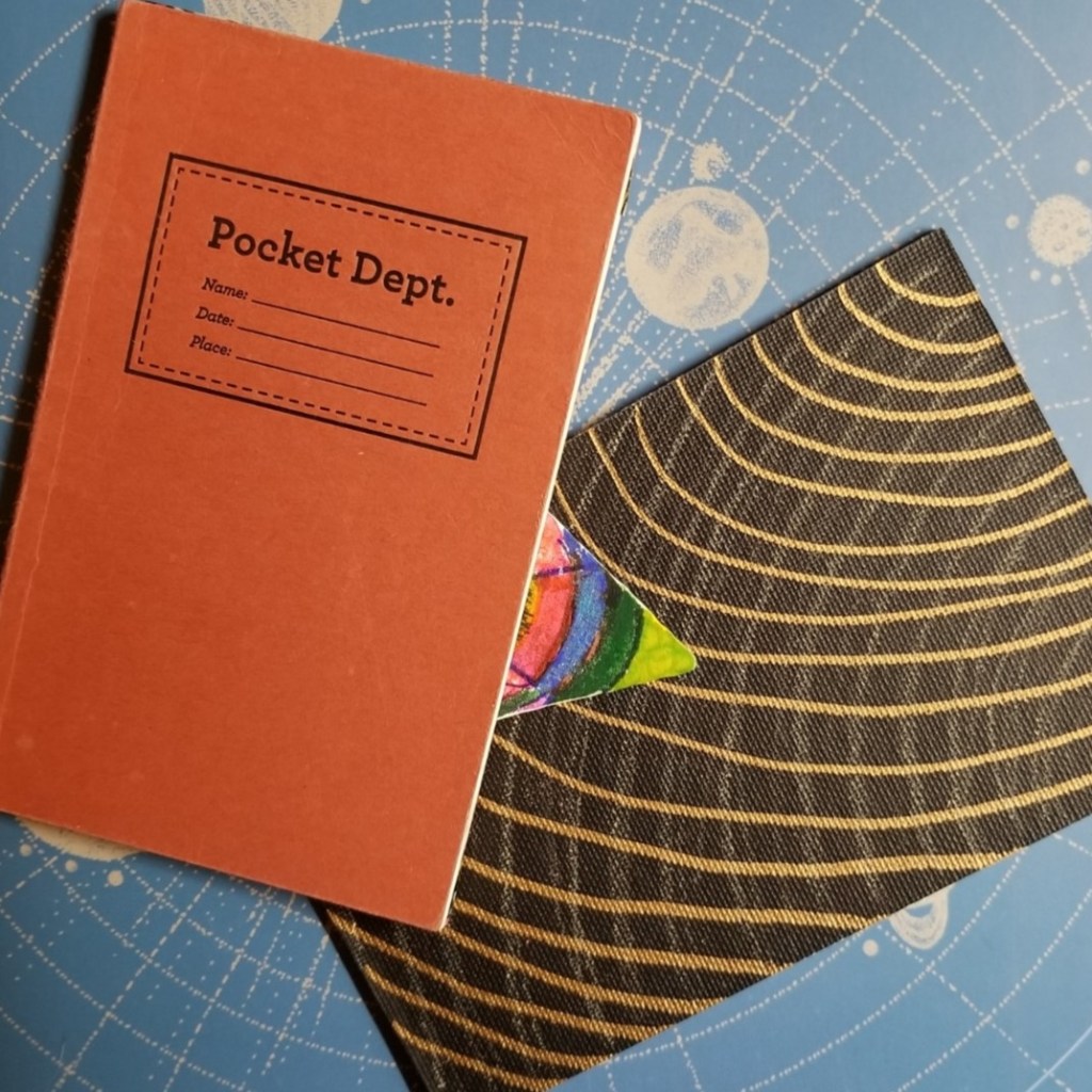 A small notebook with the words "Pocket Dept." on it. A small triangle coloring visible of a larger piece. A black postcard with two sets of curves drawn in gold. All atop a file folder with planets and orbits drawn on it.