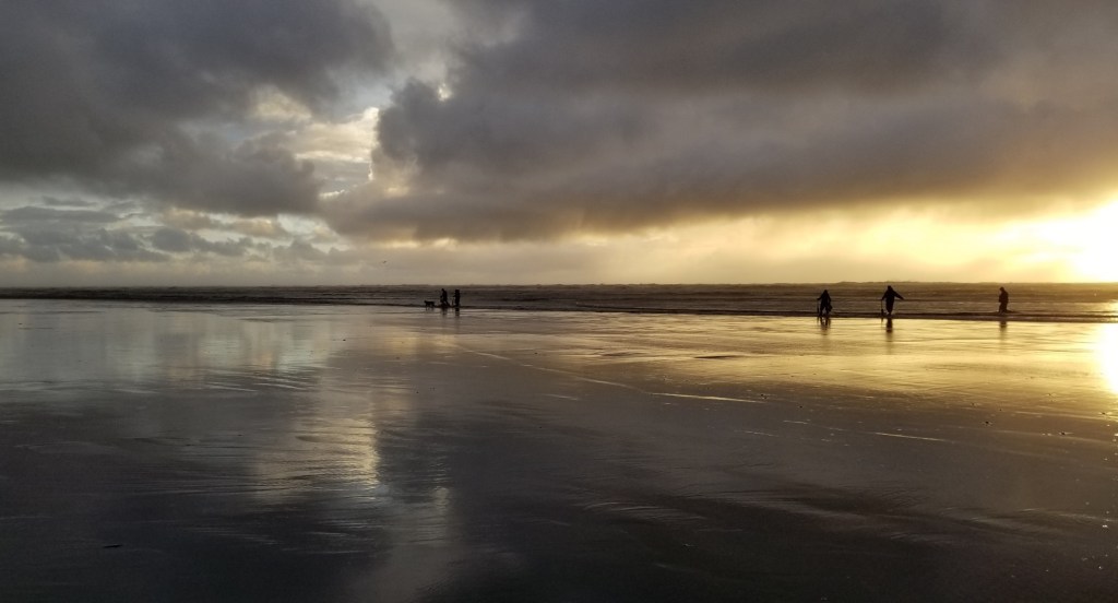 A cloudy sky with hints of blue on the left, and a setting sun on the right. The water on the sand reflects the sky above as people (seen only in dark shadows) walk beside the water.