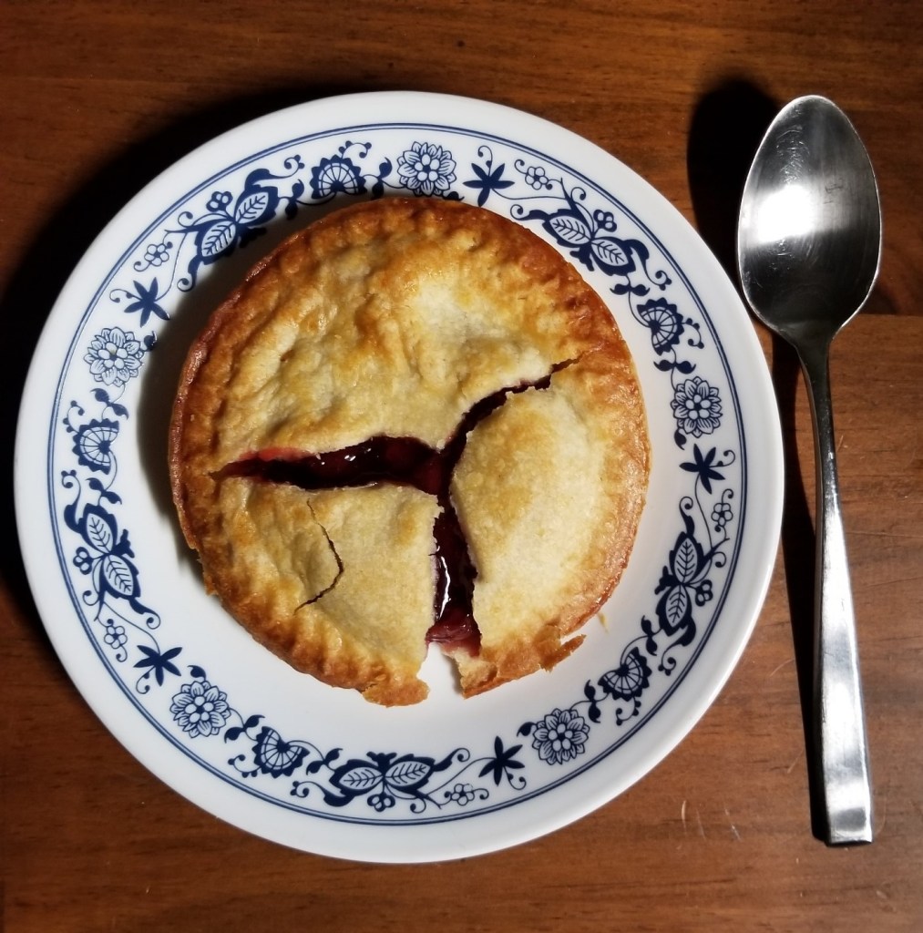 A mini/personal cherry pie on a dessert plate, with a spoon to the right of it.
